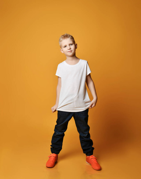 Calm, smart young schoolboy kid in blue jeans and sneakers stands showing his white blank t-shirt - Foto, Imagem
