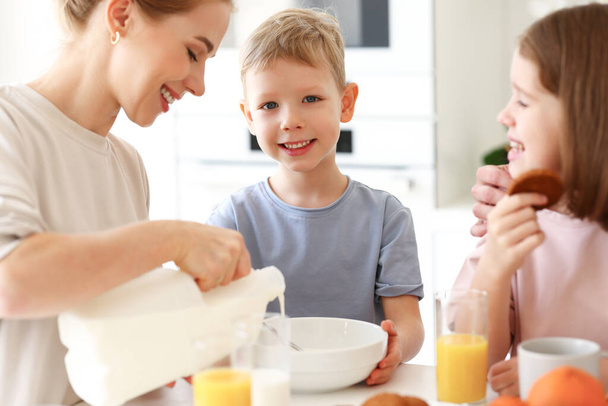 Young positive mom pouring fresh milk from bottle in glass while having healthy breakfast with two cute kids, daughter and son. Happy family of three spending time in kitchen while eating morning meal - Photo, Image