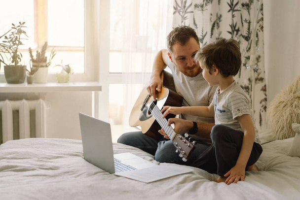 Πατέρας και γιος μαθαίνουν να παίζουν ακουστική κιθάρα σε ένα διαδικτυακό μάθημα. Ελεύθερος χρόνος να περάσω με το γιο μου στο σπίτι, μαθαίνοντας του να παίζει κιθάρα. Ευτυχισμένη ημέρα του πατέρα - Φωτογραφία, εικόνα