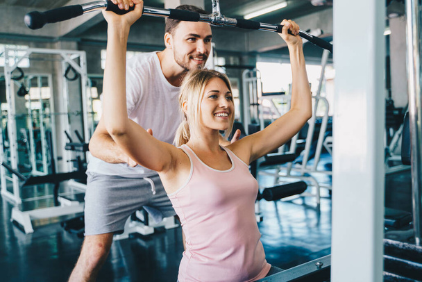Cheerful young woman wearing pink sports bra while doing chin-up exercise with trainer man in fitness gym - Foto, afbeelding