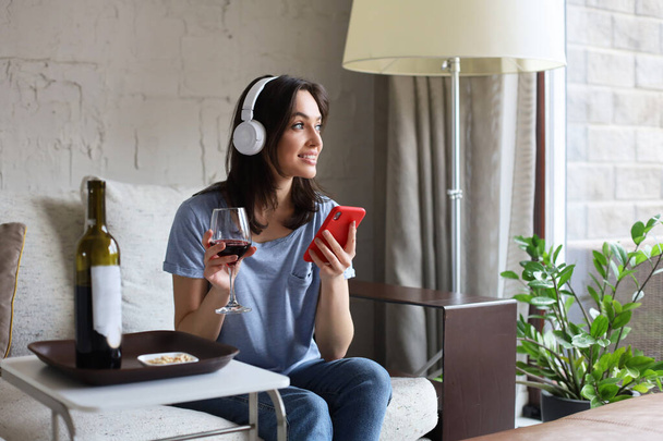 Pretty girl using her smartphone on couch at home in the living room. Listening music, drinking red wine, relaxation after a hard week at work - Foto, Imagen