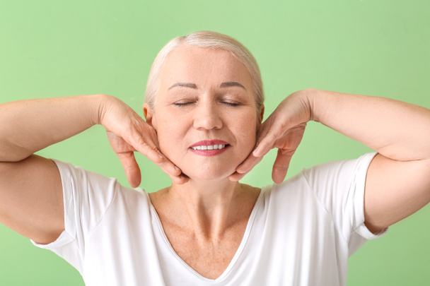 Mature woman giving herself face massage on color background - Photo, Image