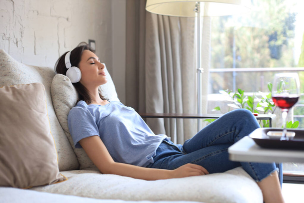 Beautiful brunette woman in earphones sitting on sofa listening to music with closed eyes - Фото, изображение