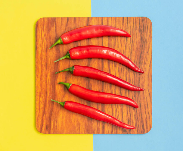 červené chilli papričky na hnědém dřevěném talíři, ležet na pastelově žluté a modré pozadí, ploché ležel čtvercový koncept - Fotografie, Obrázek
