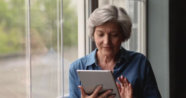 Smiling 60s older woman using tablet, standing near window. - Materiaali, video