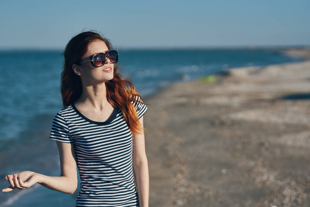 vrouw die in de zomer op het strand loopt met een bril en een gestreept t-shirt aan - Foto, afbeelding
