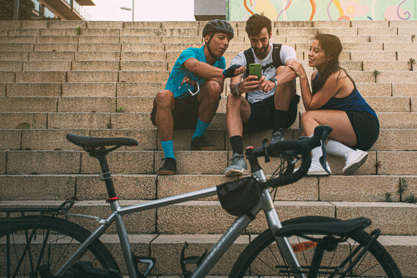 Eine Gruppe fröhlicher Freunde draußen spielt mit einem Mobiltelefon und sitzt in Sportkleidung auf einer Treppe der Stadt, vor sich ein Fahrrad. Hintergrund mit Copyspace. Technologiekonzept. Sportkonzept. Outdoorkonzept. Freizeitkonzept. Vertica - Foto, Bild