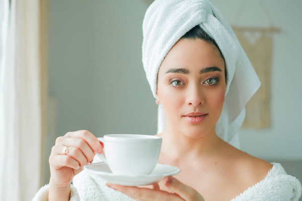 シャワーの後、頭にタオルをつけたバスローブの若い女性がコーヒーを飲み、窓から外を眺めます。朝だ。朝食. - 写真・画像