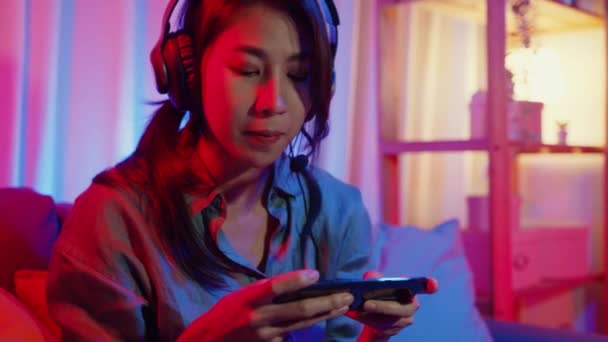 Ευτυχισμένη Ασία gamer κορίτσι φορούν ακουστικά διαγωνισμό βίντεο παιχνίδι σε απευθείας σύνδεση με smartphone ενθουσιασμένοι μιλάμε με φίλο καθίσει στον καναπέ σε πολύχρωμα φώτα νέον σαλόνι στο σπίτι, Αρχική καραντίνα έννοια δραστηριότητας. - Πλάνα, βίντεο