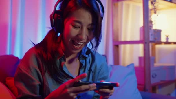 Happy azië meisje gamer dragen hoofdtelefoon concurrentie video game online met smartphone opgewonden praten met een vriend zitten op de bank in kleurrijke neon lichten woonkamer thuis, Home quarantaine activiteit concept. - Video