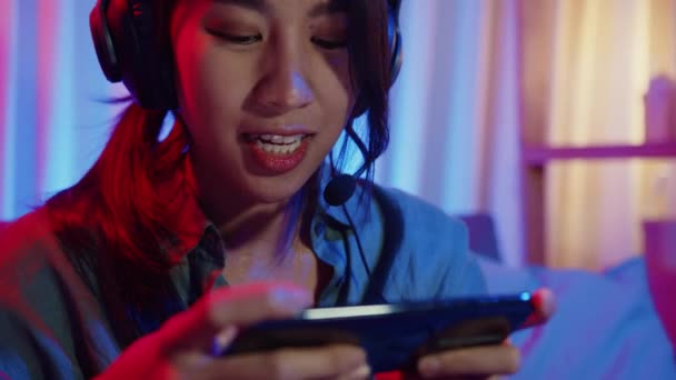 Glückliche asiatische Mädchen Gamer tragen Kopfhörer Wettbewerb Videospiel online mit Smartphone aufgeregt reden mit Freund sitzen auf der Couch in bunten Neonlichtern Wohnzimmer zu Hause, Home Quarantäne Aktivitätskonzept. - Filmmaterial, Video