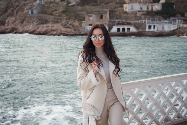 A carefree Caucasian woman in beige clothing enjoying the view of the sea on a warm, windy day. a woman wearing stylish sunglasses enjoying a beautiful winter day and breathing in the sea air - Photo, Image