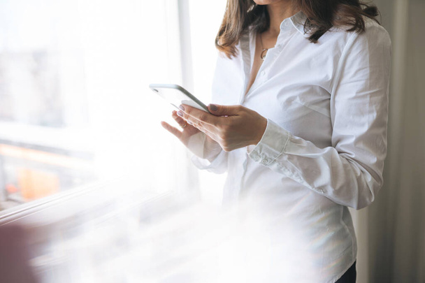 Crop photo of young brunette woman in white shirt using mobile phone in hands near window - Foto, afbeelding