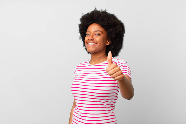 Αφρο μαύρη γυναίκα αισθάνεται υπερήφανη, ανέμελη, με αυτοπεποίθηση και ευτυχισμένη, χαμογελώντας θετικά με τους αντίχειρες επάνω - Φωτογραφία, εικόνα