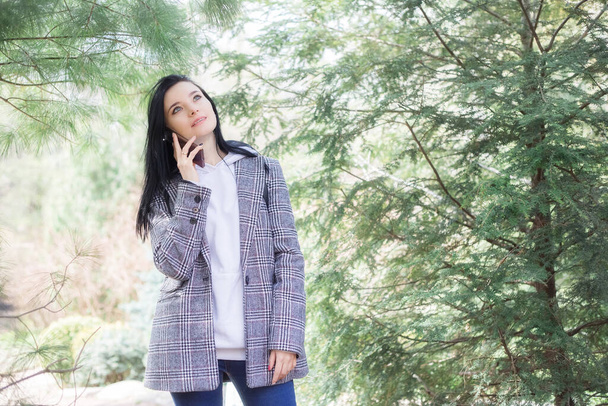 Portrait of a cute young happy brunette pretty woman in a plaid coat walking outdoors in park in beautiful spring day using mobile phone. - Foto, Imagen