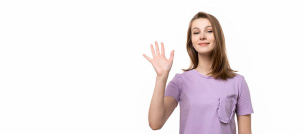 Дружня дівчина каже привіт, посміхається радісно і доброзичливо, махає рукою, показуючи п'ять пальців, доброзичливий вітальний жест. Студійний знімок, білий фон
 - Фото, зображення
