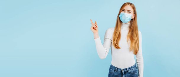 Image of happy young woman wearing medical protective mask on face pointing finger to the side at copy space on isolated blue background - Foto, immagini