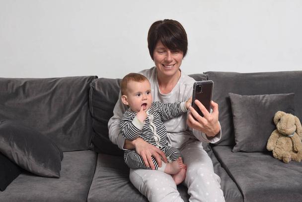 ソファに座っている母親と息子母親はスマートフォンを赤ちゃんに見せます - 写真・画像