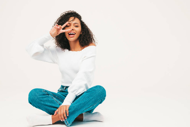 Beautiful black woman with afro curls hairstyle.Smiling model in sweater and trendy jeans clothes. Sexy carefree female posing on white background in studio. Tanned and cheerful.Shows peace sign - Fotografie, Obrázek