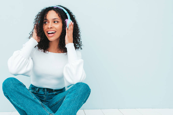 Beautiful black woman with afro curls hairstyle.Smiling model in sweater and jeans.Sexy carefree female listening music in wireless headphones.Posing in studio near light blue wall - Foto, immagini