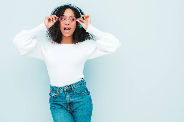 Beautiful black woman with afro curls hairstyle.Smiling model in sweater and jeans.Sexy carefree female listening music in wireless headphones.Posing in studio near light blue wall.Shows peace sign - Foto, afbeelding