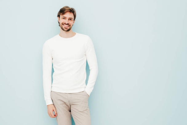カジュアルな白いセーターとズボンを着てハンサムな笑顔ヒップスター腰掛け性的ビジネスマンモデルの肖像画。ファッションスタイリッシュな男ポルノのスタジオでライトブルーの壁に対して - 写真・画像