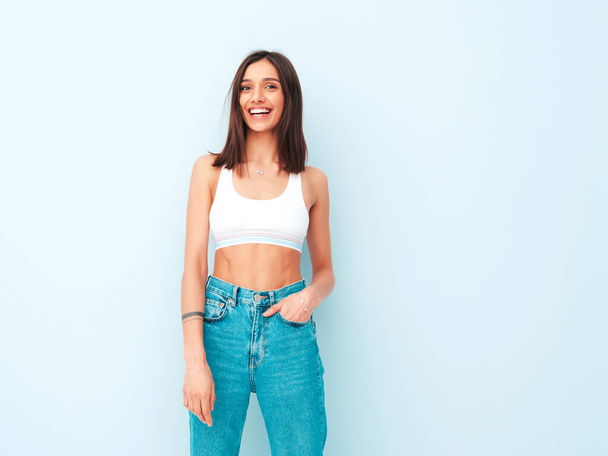 Beautiful smiling woman dressed in white jersey top shirt and jeans. Sexy carefree cheerful model enjoying her morning. Adorable and positive female posing near light blue wall in studio - Photo, image