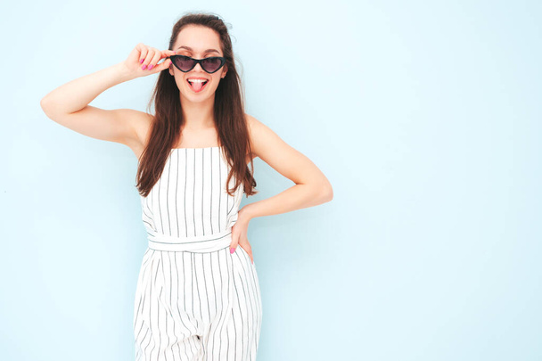Portrait of young beautiful smiling female in trendy summer hipster overalls clothes. Sexy carefree woman posing near light blue wall in studio. Positive model having fun indoors.Shows tongue  - Φωτογραφία, εικόνα