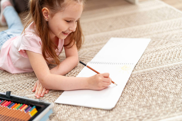 Little cute Caucasian girl child, drawing in an album with colored pencils, lying on floor. hardworking child does homework. The child's hobby is drawing. new modern set for drawing and creativity - Foto, Imagem