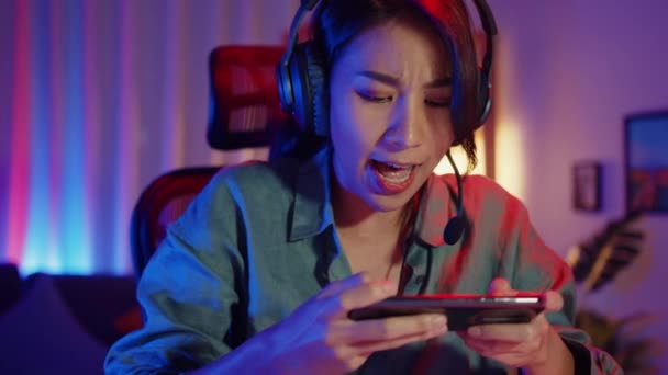 Ευτυχισμένη Ασία gamer κορίτσι φορούν ακουστικά διαγωνισμό παίζουν βιντεοπαιχνίδι σε απευθείας σύνδεση με smartphone πολύχρωμα φώτα νέον στο σαλόνι στο σπίτι. Esport streaming παιχνίδι σε απευθείας σύνδεση, Αρχική καραντίνα έννοια δραστηριότητας. - Πλάνα, βίντεο