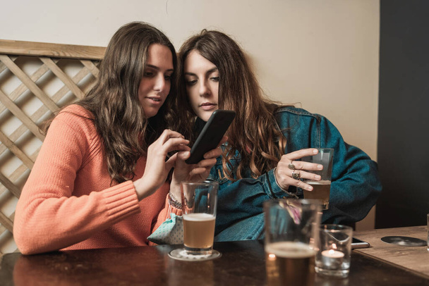 Duas jovens amigas compartilhando a tela do smartphone bebendo cervejas sentadas na mesa dos pubs. Juventude usando álcool e conceito de tecnologia. - Foto, Imagem