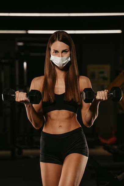 Una mujer en forma en una máscara facial para evitar la propagación del coronavirus está haciendo rizos de bíceps con mancuernas. Una chica deportiva con una máscara quirúrgica posando durante el entrenamiento de brazos en un gimnasio. - Foto, imagen