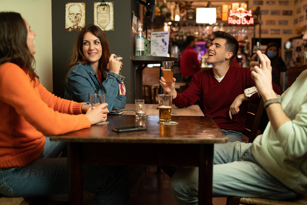 Złoczyńcy żartują razem pijąc piwo i wznosząc toast w pubie. Koncepcja młodych ludzi zbierających się i bawiących w nocy - Zdjęcie, obraz