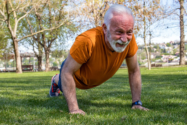 Portret starszego mężczyzny ćwiczącego, robiącego pompki w parku. Starszy mężczyzna w dobrej formie z silnymi ramionami. - Zdjęcie, obraz