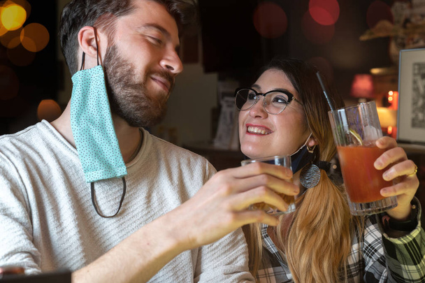 Couple de jeunes gens avec masque facial abaissé cliquetis lunettes ensemble. Un homme et une femme s'amusent à trinquer pendant le coronavirus nouveau temps normal. - Photo, image