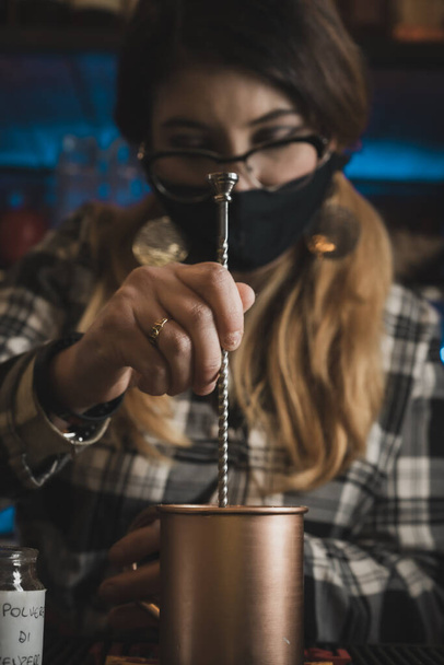 bartender pounds the ingredients for the preparation of Moscow mule cocktail  with a mortar inside the characteristic mug wearing the protective mask due to the coronavirus pandemic - Zdjęcie, obraz