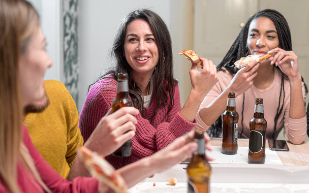 Young people at home having fun taking a pizza party together. Multiracial millennial generation friends drinking beers and eating slices of pizza - Photo, Image