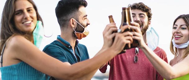 Group of young friends having fun drinking and clinking with beers after the coronavirus lockdown. Happy people with protective masks toasting with beer bottles together. - Photo, Image
