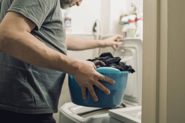 Hombre adulto lavando ropa usando una lavadora de carga superior. Primer plano de la mano sosteniendo un lavabo que contiene ropa sucia - Foto, imagen