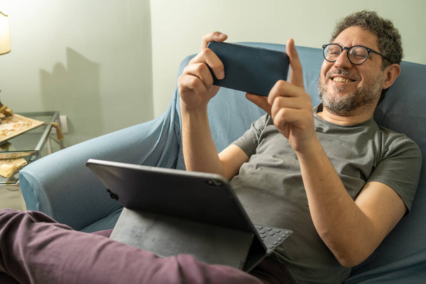 Hombre adulto de mediana edad escuchando música o haciendo una videollamada en línea en el sofá con auriculares inalámbricos - Foto, Imagen