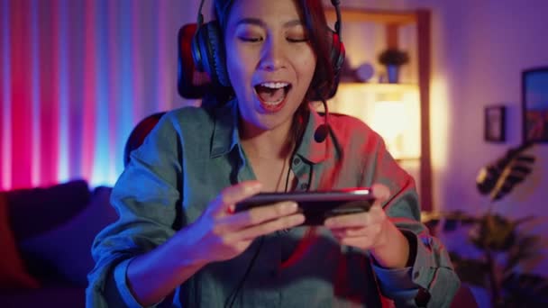 Happy ASia девушка геймер носить наушники конкуренции играть в видеоигры онлайн со смартфоном красочные неоновые огни в гостиной дома. Онлайн-трансляция игры Esporte, Концепция домашнего карантина. - Кадры, видео