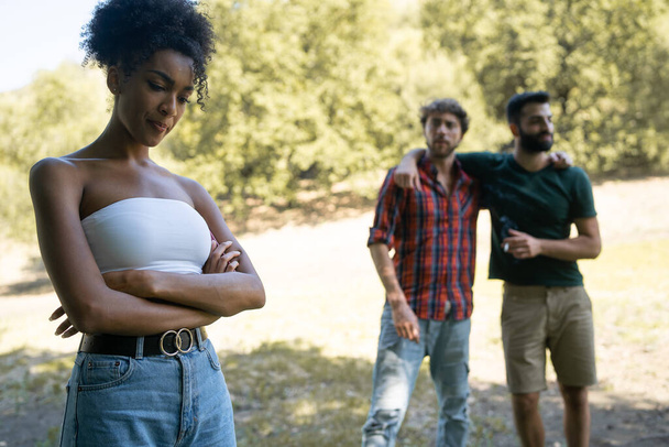 A girl of African descent with folded arms is bullied by two young men in an outdoor park. Concept of bullying, racism among young people. - Photo, Image