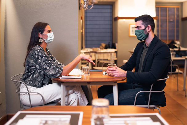 Pár rozhovorů sedí u stolu v restauraci, nosí masky, v novém normálu po pandemii koronaviru. COVID-19 koncept životního stylu. - Fotografie, Obrázek