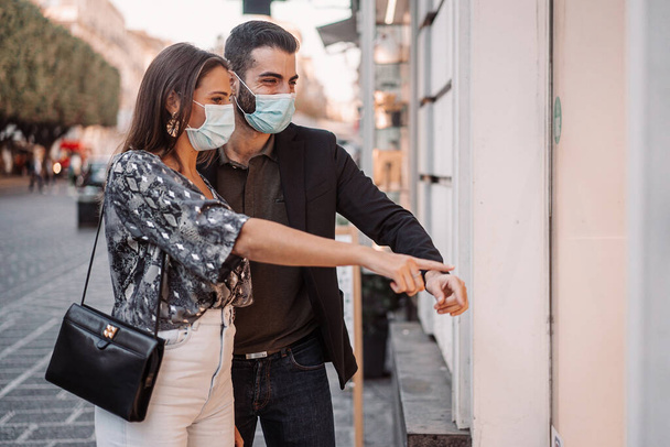 市内のショップウィンドウを一緒にいくつかの商品を指して見て顔のマスクを歩く若い人たちのカップル。コロナウイルスの拡散中に新しい普通の言葉で何かをする人々の概念. - 写真・画像