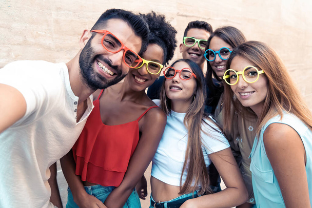 Cheerful multiethnic group of millennial generation z friends wearing funny glasses embracing arms on shoulders smiling and having fun isolated against a white wall. Diversity lifestyle concept - Photo, Image