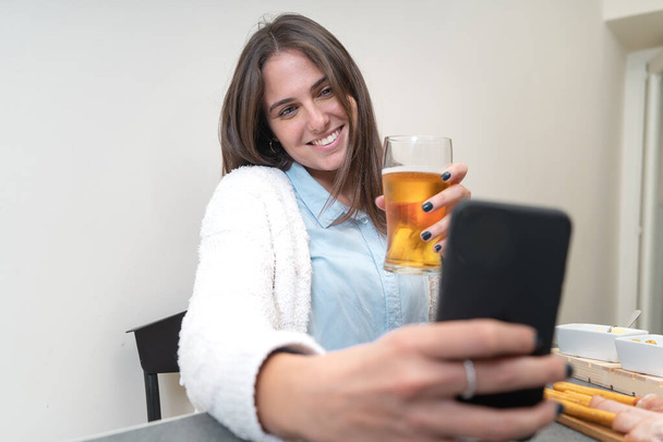 Una joven bebe cerveza durante una videollamada con su novio o amigos. Ella se sienta en una mesa en la cocina en casa. - Foto, Imagen