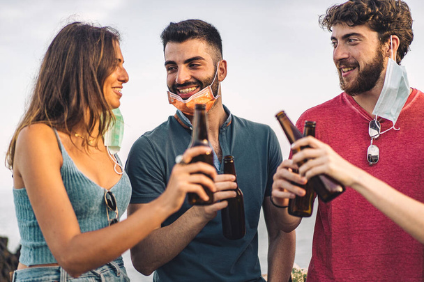 New normal lifestyle concept of young people hangout toasting with beer bottles outdoors during coronavirus breakdown wearing lowered face masks - Valokuva, kuva