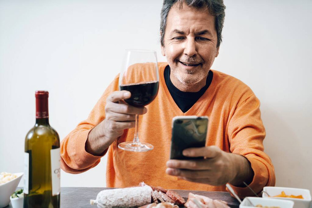 Красивый пожилой мужчина тост дома с красным вином глядя на камеру на нейтральном белом фоне - Фото, изображение