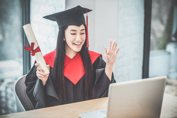Cute asian smiling graduate holding her diploma and having a video call - Fotoğraf, Görsel