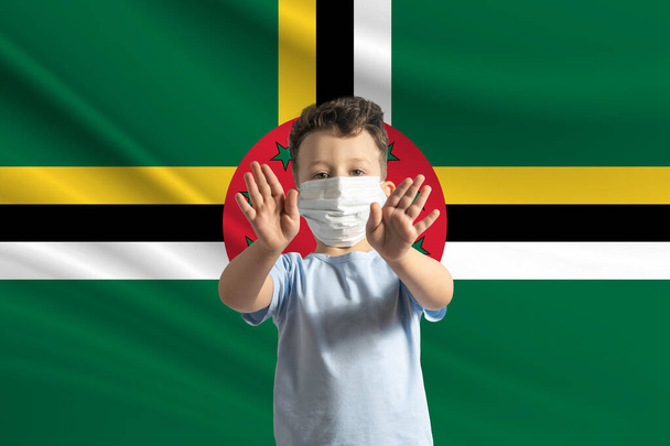 Μικρό λευκό αγόρι με προστατευτική μάσκα στο φόντο της σημαίας της Ντομίνικα κάνει στοπ με τα χέρια του, μένει σπίτι Ντομίνικα. - Φωτογραφία, εικόνα